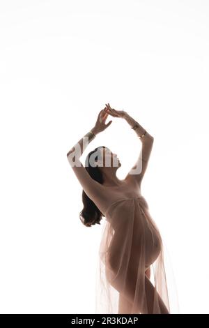 future mother in golden bracelets and transparent chiffon cloth posing with closed eyes and raised hands isolated on white background, maternity style Stock Photo