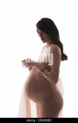 charming and graceful brunette pregnant woman in golden bracelets and delicate chiffon draping standing isolated on white background, maternity fashio Stock Photo