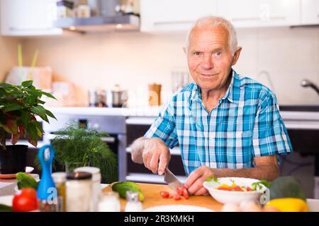 elderly man cuts vegetables for salad at the table in the kitchen Stock Photo