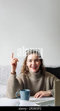 Eureka Excited female entrepreneur with successful business idea, sitting at kitchen table with laptop and showing index finger, ready to make a Stock Photo