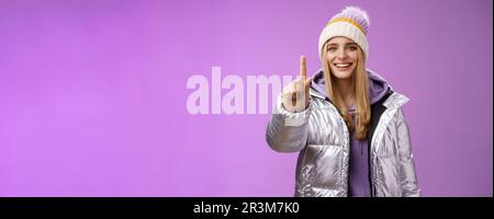 Excited cheerful fair-haired european girl in winter hat silver shiny jacket show number one index fingers give suggestion advic Stock Photo
