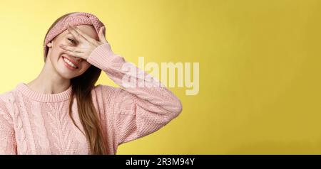 Blushing cute girl feeling positive, smiling happy joyful covering eyes with palm peeking through fingers delighted, playful, ti Stock Photo