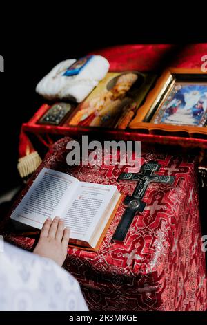 Hand of priest holds opened Holy Bible on the lectern near cross and icons for rite in Orthodox Ukrainian church. Top view. Stock Photo