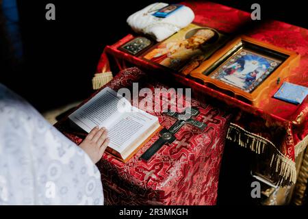 Hand of priest holds opened Holy Bible on the lectern near cross and icons for rite in Orthodox Ukrainian church. Top view. Stock Photo