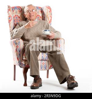 Senior Citizens: Tea and Biscuits. A welcome break for a smiling man in an armchair. Full length portrait, isolated on white. From a series. Stock Photo