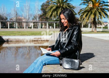 Side view of smiling young Hispanic student in black leather jacket and jeans sitting on a wooden frame with a handbag and looking down as she reads a Stock Photo