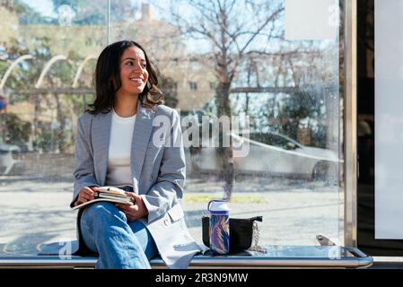 Positive young Hispanic female student sitting cross legged on bench with books water bottle at city bus stop with glass walls and smiling while looki Stock Photo