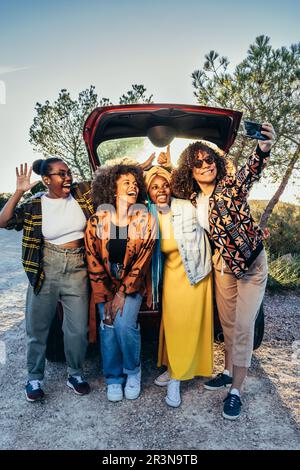 Front view of group of cheerful African American female friends looking at screen of mobile phone and taking selfie while standing and leaning on park Stock Photo