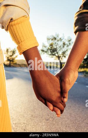 Anonymous African American female friends in casual clothes holding hands and standing on city street during daytime against blurred green tree Stock Photo