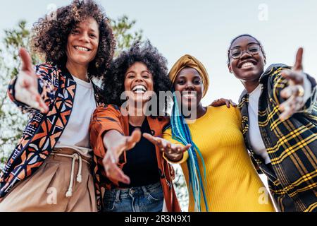 From below group of happy multiethnic female friends in stylish clothes smiling and looking at camera while standing under bright blue sky in park at Stock Photo