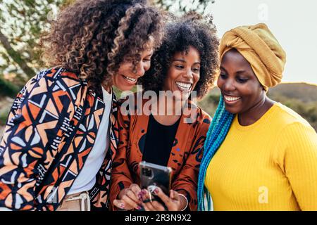 Young cheerful multiracial female friends in stylish clothes smiling and browsing smartphone while standing together Stock Photo