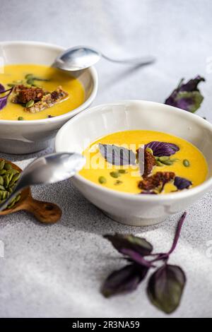 Front view of cropped bowls with pumpkin cream soup with basil herb, rye bread and seeds on blurred grey background with leaves Stock Photo
