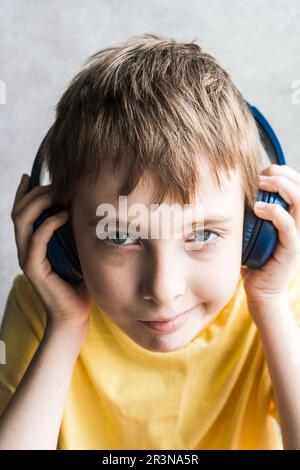 Front view of smiling boy with brown hair wearing yellow t-shirt and touching headphones while listening to music and looking at camera against blurre Stock Photo