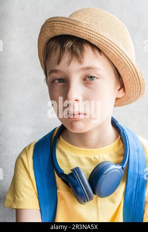 Portrait of serious boy with blue eyes and brown hair wearing yellow t-shirt and hat with blue bag pack and headphones on neck against blurred backgro Stock Photo