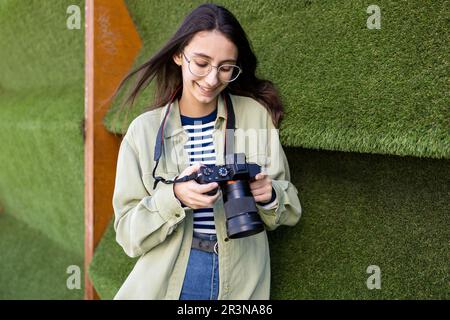 Young female photographer in casual clothes smiling and looking at photo camera while standing near green wall Stock Photo