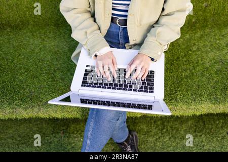 From above unrecognizable young remote worker sitting on a grassy knoll and browsing her laptop while working in a park during the workday Stock Photo