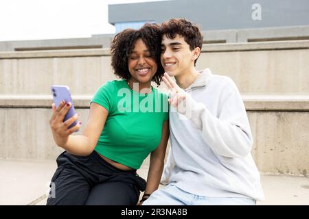 Cheerful young multiracial friends in casual clothes taking selfie on smartphone while sitting on concrete slab of building in daylight outdoors Stock Photo