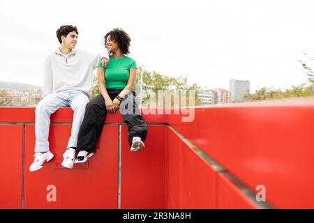 Confident multiracial couple sitting on red fence and looking at each other while male leaning on female's shoulder and resting in bright sunlight aga Stock Photo