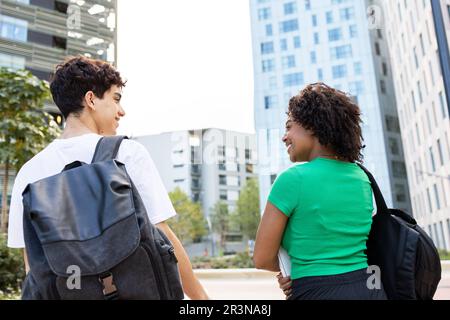 Back view of smiling young male and female with curly hair looking at each other while walking with backpacks on college campus road with buildings in Stock Photo