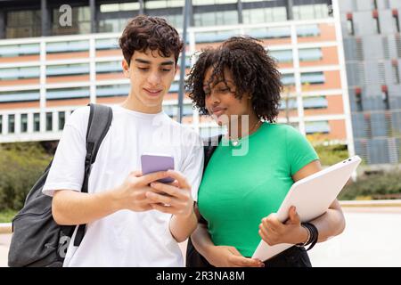 Positive young multiracial male and female students with backpacks looking at screen of mobile phone while standing together in daylight near modern b Stock Photo
