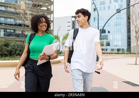 Positive multiracial male and female students with back packs walking in daylight on walkway and looking at each other while smiling and speaking in u Stock Photo