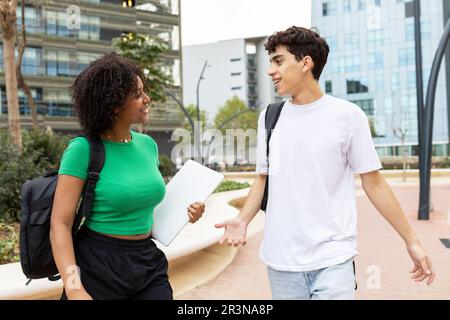 Positive multiracial male and female students with back packs standing in daylight on walkway and looking at each other while smiling and speaking in Stock Photo