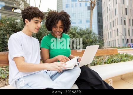 Concentrated young students sitting together on cement border with legs crossed in daylight while reading book against green trees Stock Photo