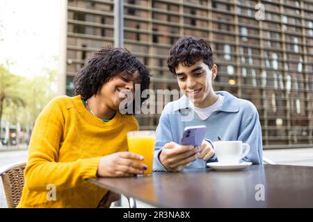 Young multiracial male and female friends smiling and looking at screen of mobile phone while sitting in daylight at table with orange juice glass and Stock Photo