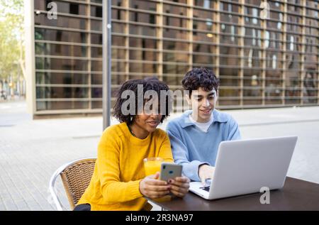 Young multiracial male and female friends smiling and looking at laptop and mobile phone while sitting in daylight Stock Photo