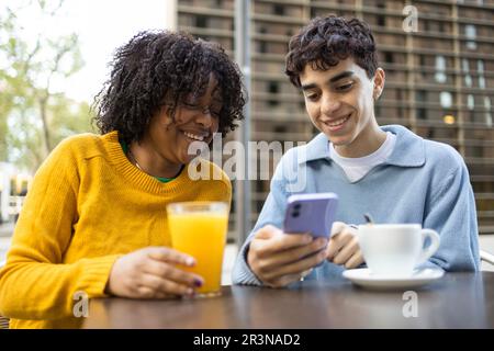Young multiracial male and female friends smiling and looking at screen of mobile phone while sitting in daylight Stock Photo