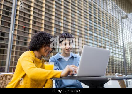 Young multiracial male and female friends smiling and looking at laptop while sitting in daylight Stock Photo