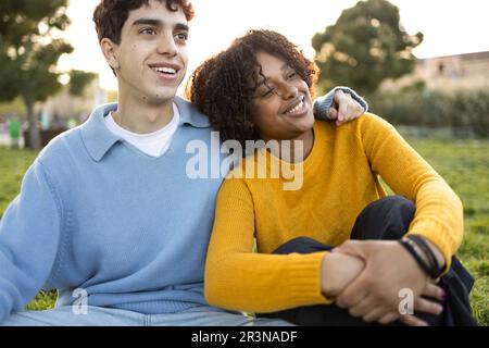 Front view of smiling multiracial young female and male friends while man leaning his arm on woman's shoulder sitting on green grass in park and looki Stock Photo
