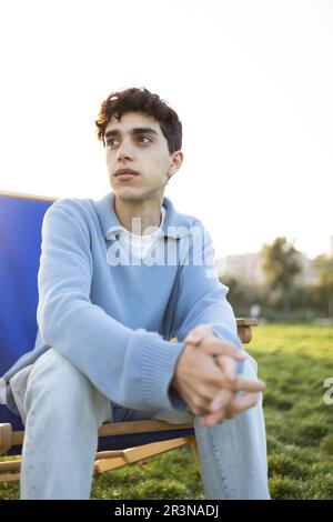Pondering young male in casual clothes sitting on deckchair with clasped hands and looking away while relaxing in green park on sunny day Stock Photo