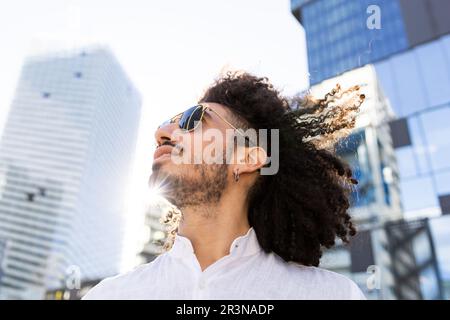 From below African American man with modern clothes, afro hairstyle and sunglasses looking at the sky in the middle of the city Stock Photo