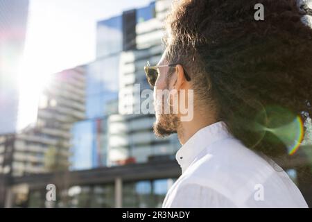 Unrecognizable African American man with modern clothes, afro hairstyle and sunglasses looking away while standing on a city street with tall building Stock Photo