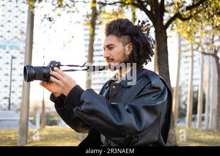 Side view of ethnic young man taking picture with a modern camera while standing on blurred background of a city street Stock Photo