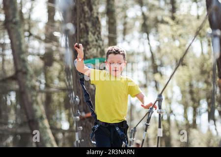 Low angle of positive preteen boy in casual clothes with safety equipment walking on rope bridge against blurred trees in woodland during summer weeke Stock Photo