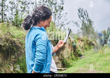 Side view of cheerful middle aged Latin American female farmer with dark hair in casual clothes smiling while messaging on mobile phone standing on gr Stock Photo