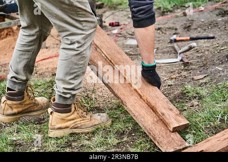 From above of crop unrecognizable male carpenter in casual wear boots and gloves carrying wooden planks during work in farm in Ecuador Stock Photo