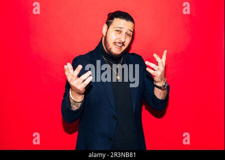Portrait of young bearded male in trendy suit showing gesture with hands up and looking at camera while standing against red background Stock Photo