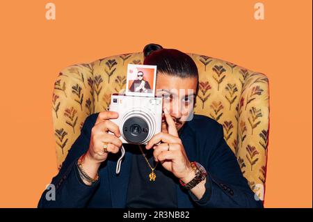 Confident young male in suit sitting in armchair and looking at camera while holding retro photo camera against orange background in studio Stock Photo