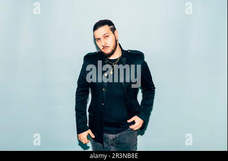 Confident bearded young male model in black jacket and denim jeans with accessories on neck looking at camera against blue background in studio Stock Photo