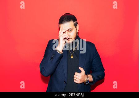 Portrait of serious young male in trendy suit with expensive wristwatch and accessories looking at camera with hand over face while standing against r Stock Photo