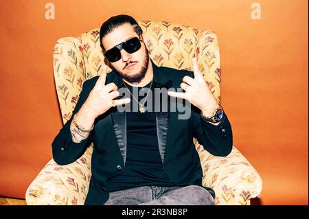 Serious young bearded male hipster wearing black jacket and sunglasses with wristwatch and necklace sitting in armchair while looking at camera and sh Stock Photo