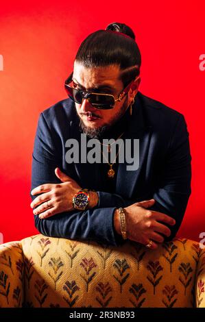 Confident bearded male model in black jacket and sunglasses with accessories looking away while leaning on armchair with crossed arms against red back Stock Photo