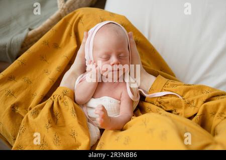 From above faceless female in yellow dress relaxing on cozy bed and holding adorable infant peacefully sleeping with closed eyes in hands Stock Photo