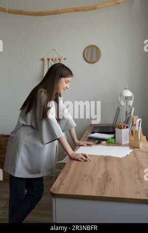 Side view of a happy and relaxed teenager standing with her hands on the table looking at the drawing with her laptop at her side Stock Photo