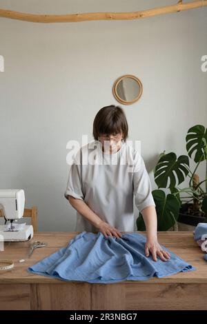 Concentrated mature female designer in eyeglasses examining blue fabric on wooden table while preparing for work on sewing machine in light atelier Stock Photo