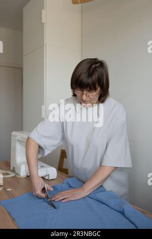 Middle age female seamstress in casual clothes and glasses standing at table while cutting fabric and working in office indoors Stock Photo