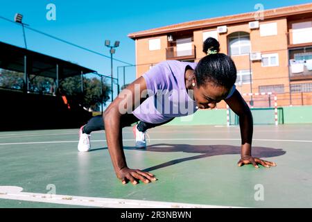 Determined adult fit African American sportswoman with dark hair in activewear doing push ups exercise while working out on outdoor sports ground unde Stock Photo